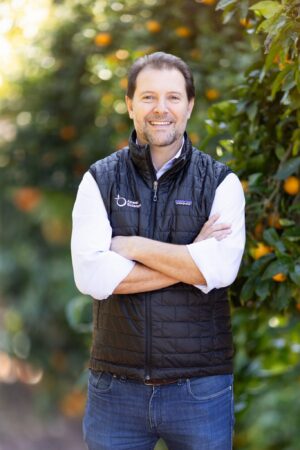 A man smiles with his arms crossed in front a fruit tree