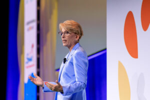Cathy Burns, dressed in a lavender blazer, addresses an audience from a stage