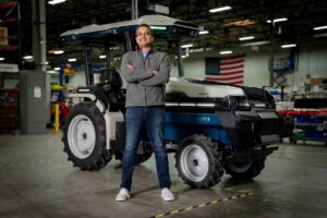 A man stands with arms crossed and a smile on his face in front of a compact, EV tractor