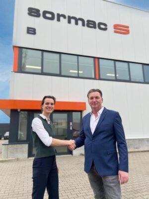 A woman and a man in business attire shake hands in front of an office building
